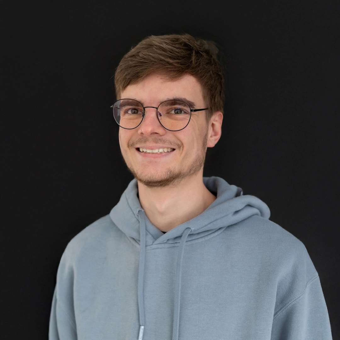 Mann mit Brille und grauem Hoodie vor schwarzem Hintergrund.